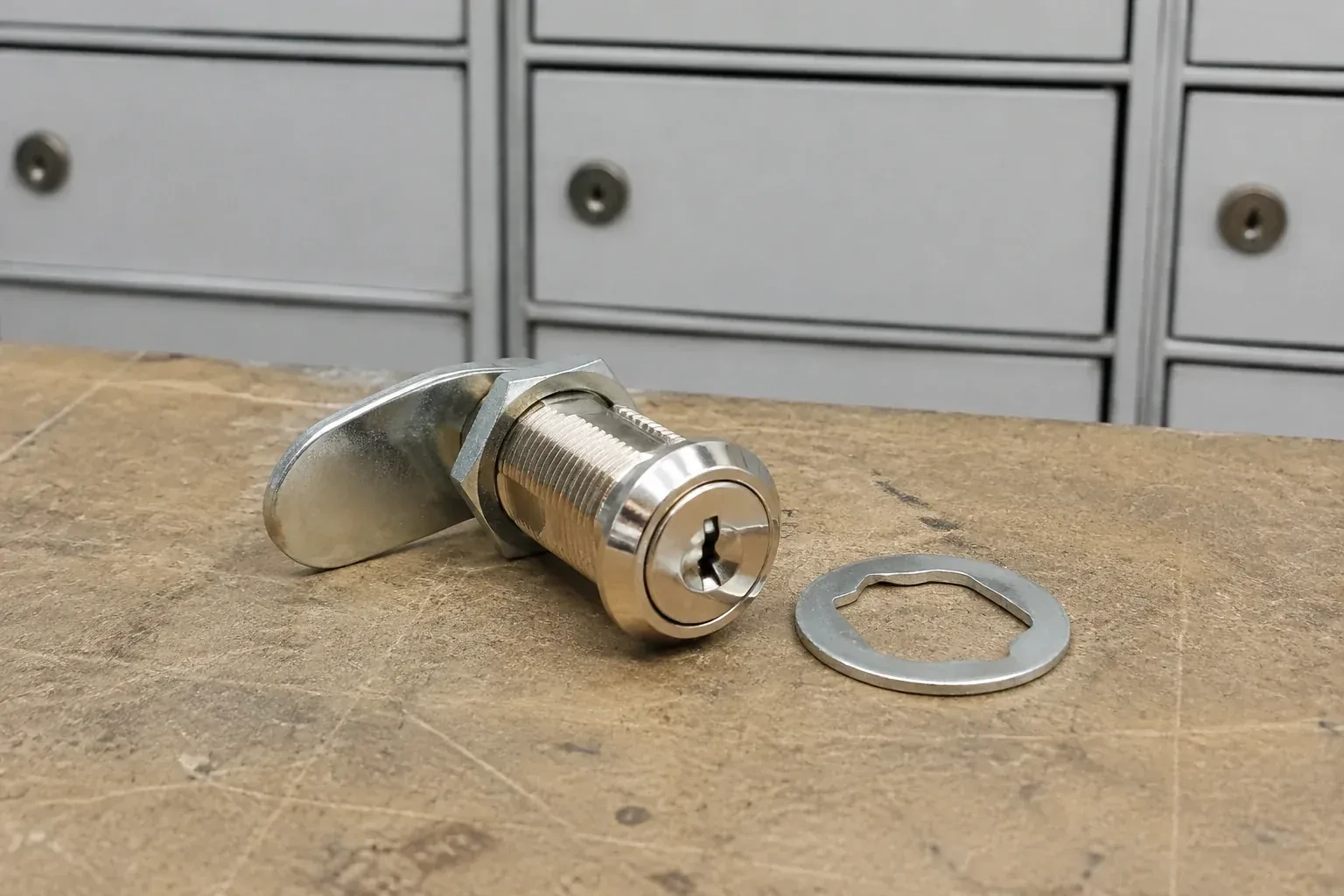 Detached threaded cam lock and mounting washer on a workbench in front of closed mail compartments. — Mailbox Lock Replacement, Cambridge